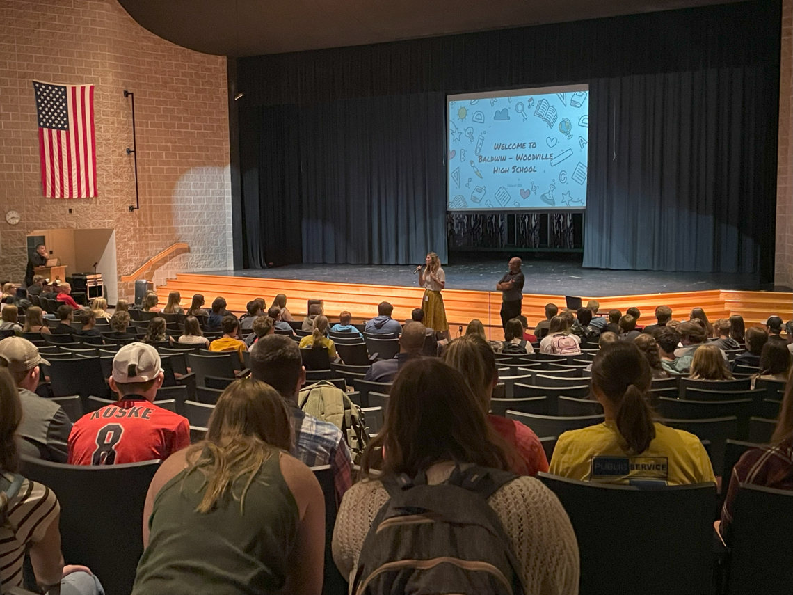 Principal and teacher addressing students in an auditorium.